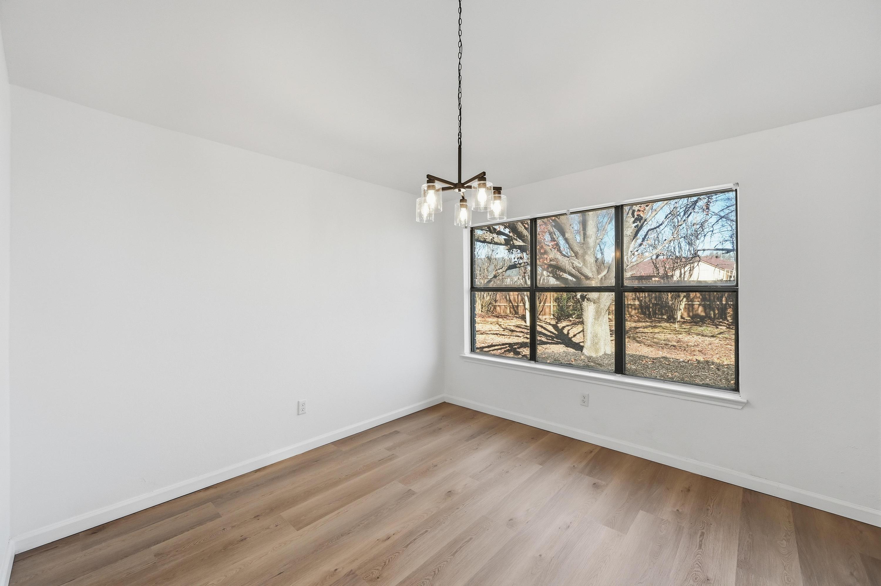 Empty Sunswept dining area with a white wall on the left, a hanging chandelier, and a large black-framed backyard window on the right